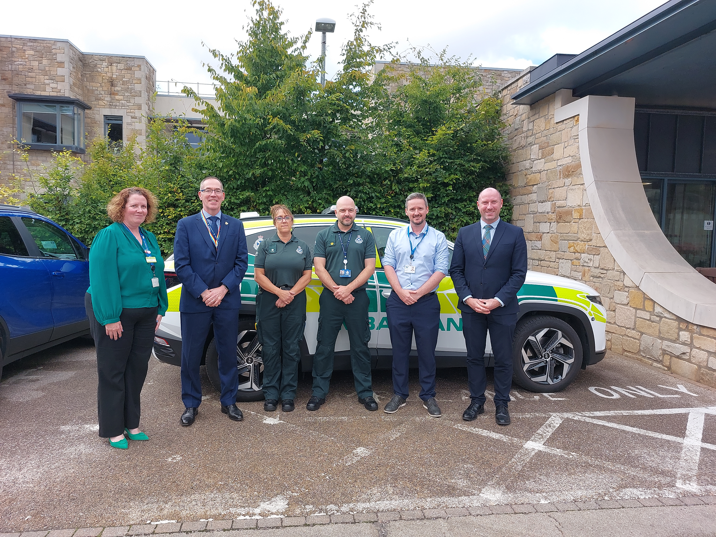 Group of 6 people standing in front of a parked ambulance car including SAS Chief Executive Michael Dickson and SG Cabinet Secretary Neil Gray.
