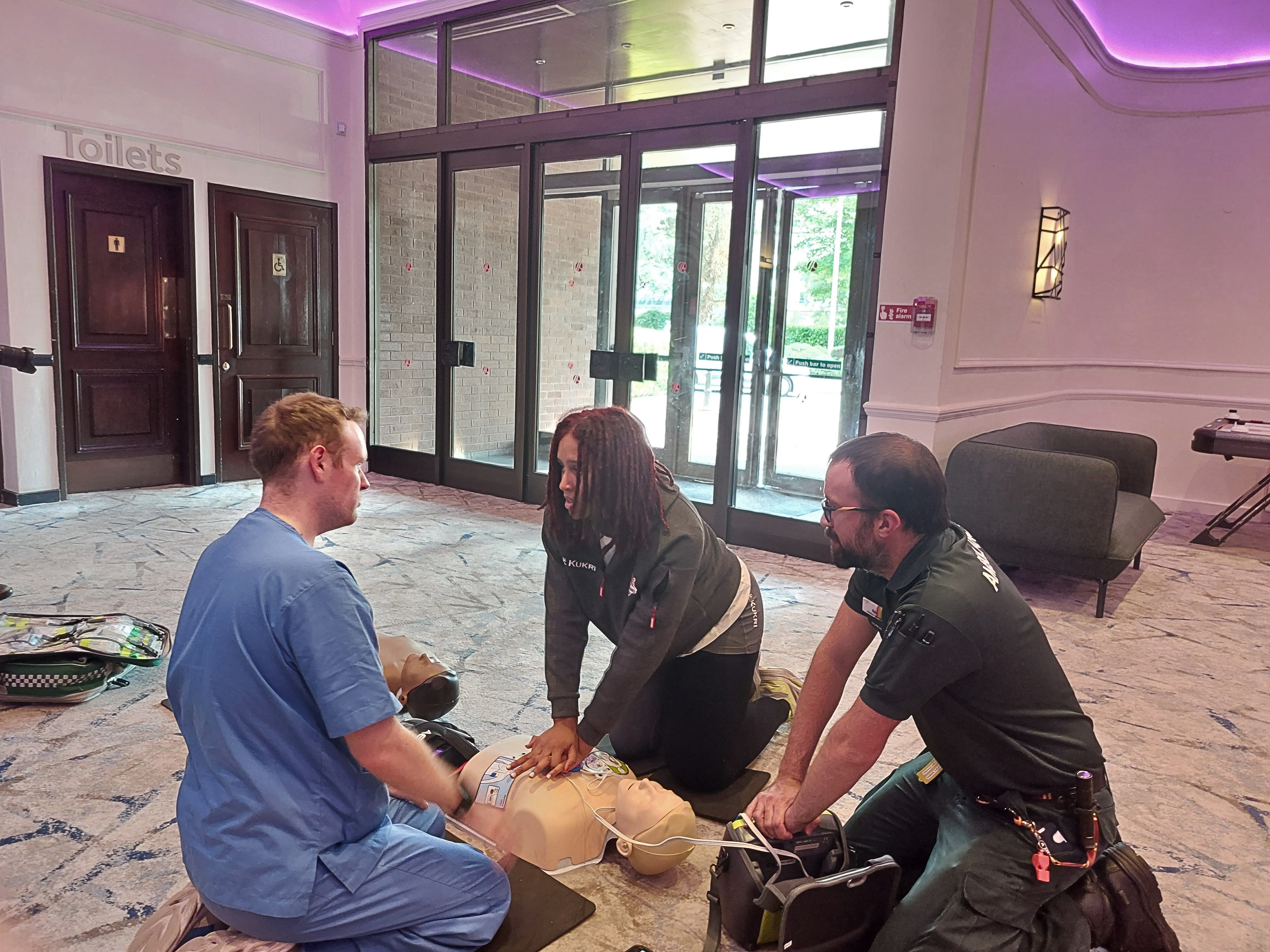 Two medical professionals instructing an athlete on how to perform CPR on a dummy.