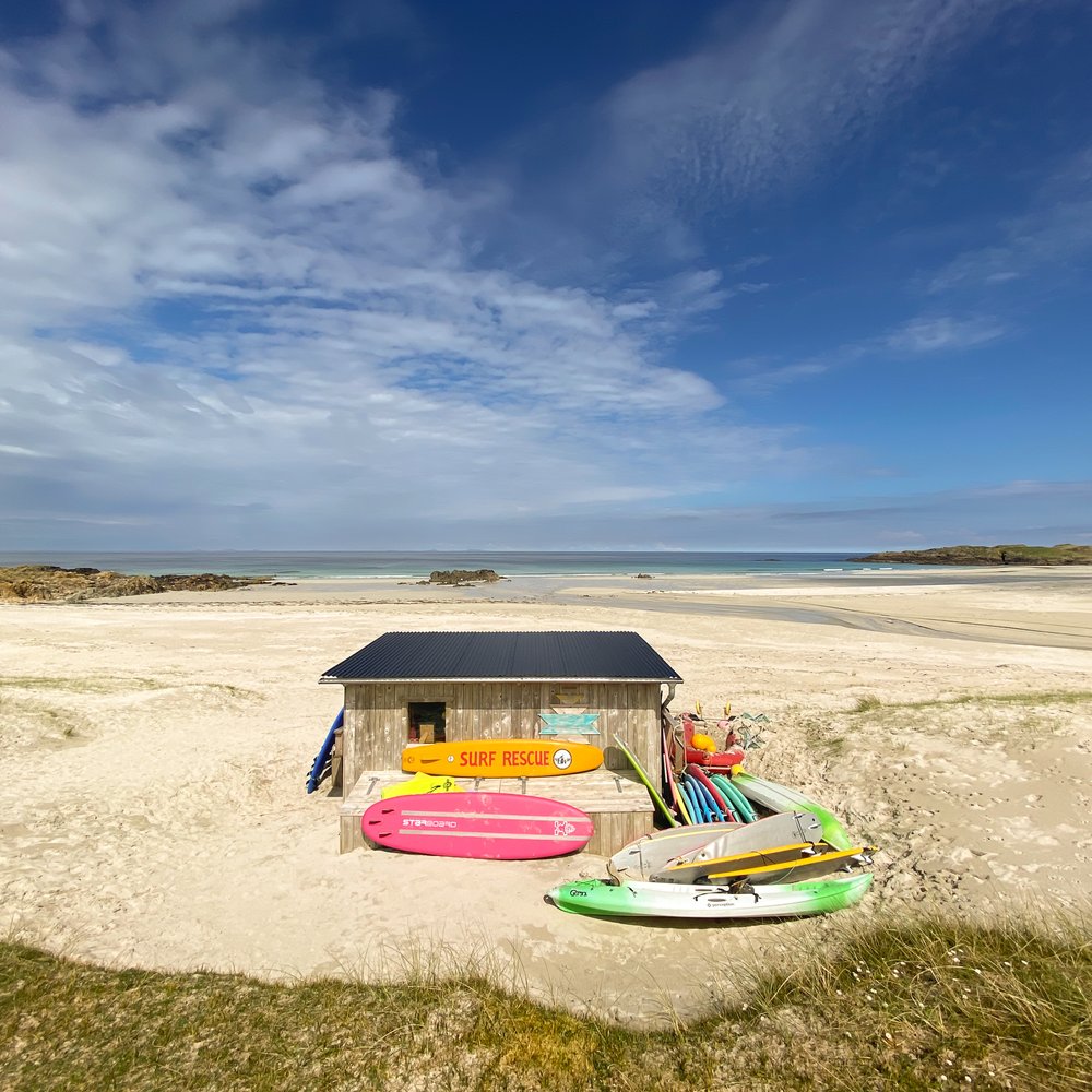 A beach shack on a beach with surf boards in front and the sea in the distance