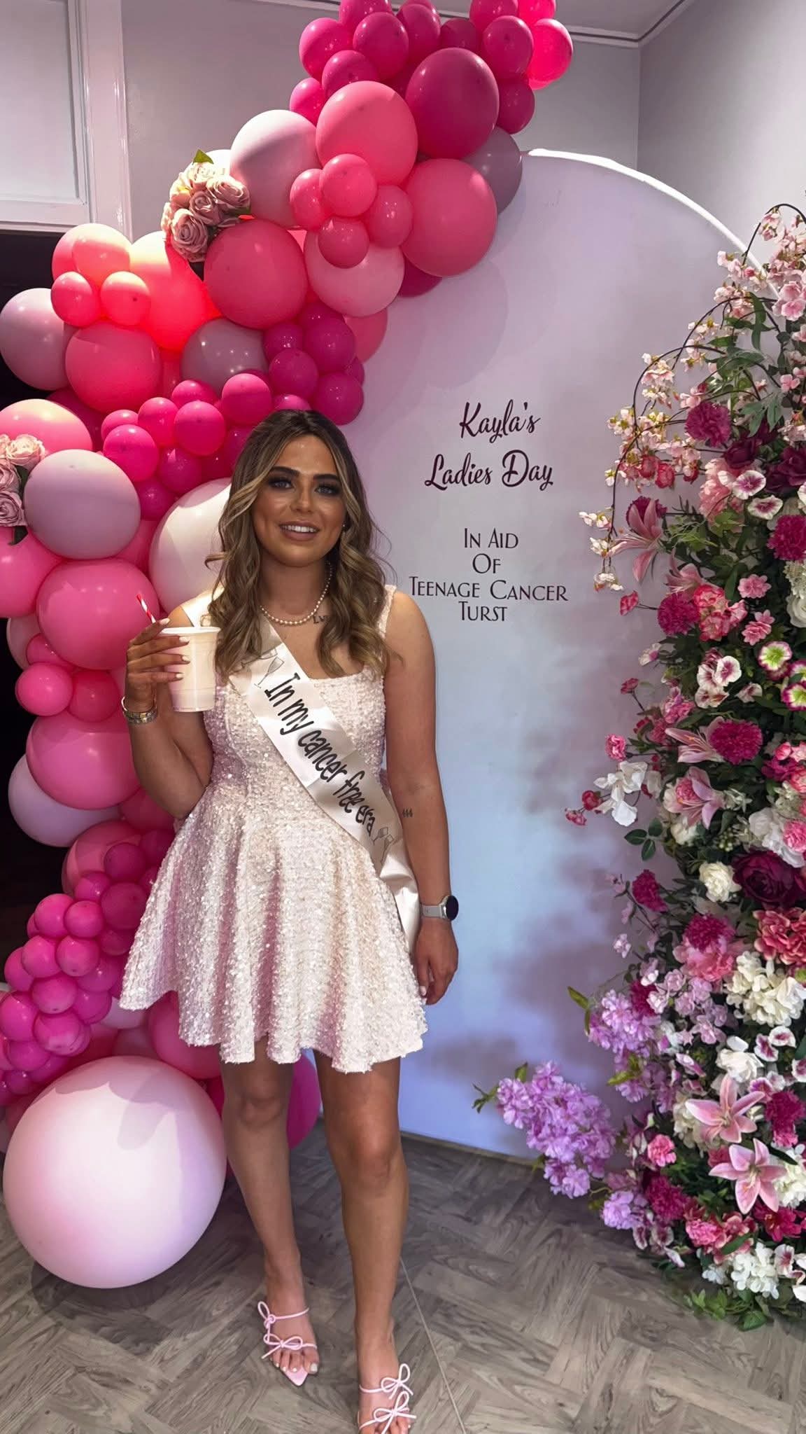 Kayla stands with a cup smiling in front of a fundraising board for the Ladies' Event