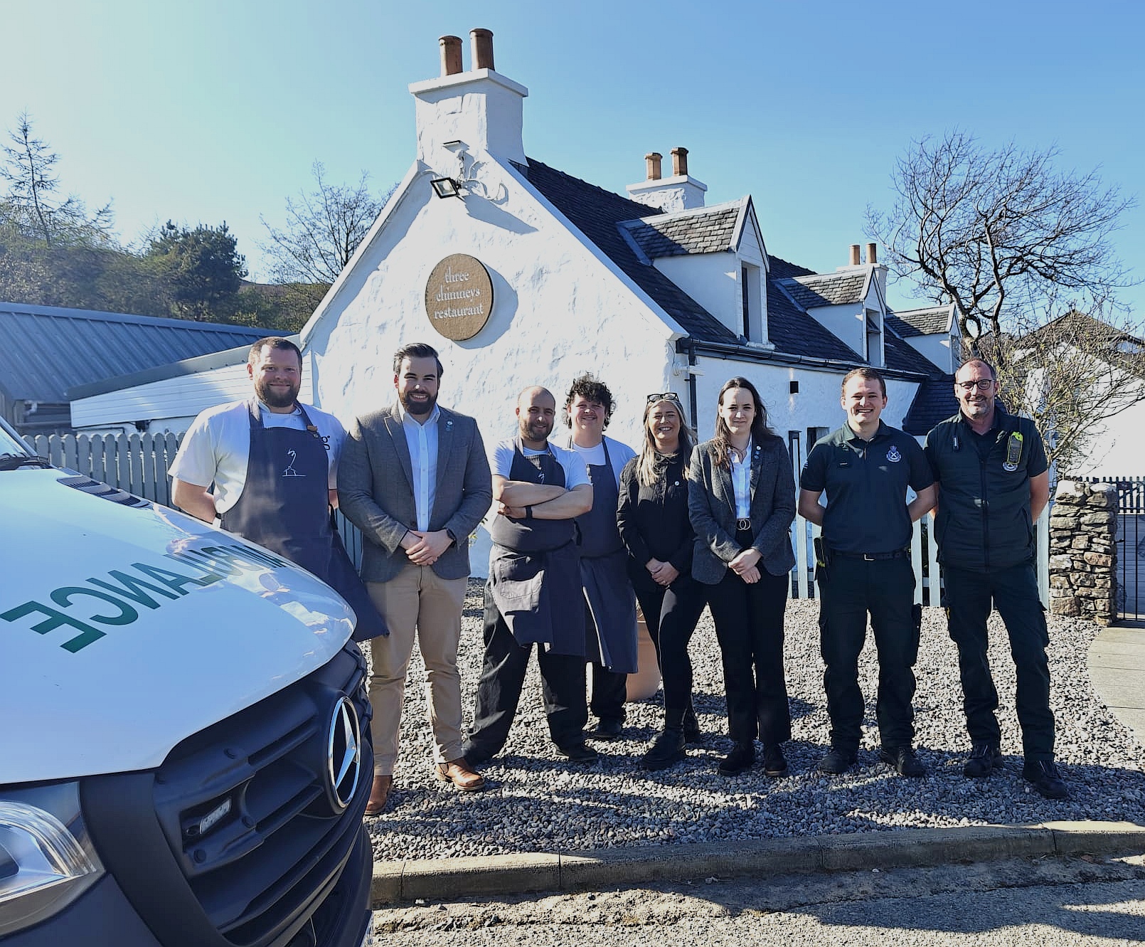 Restaurant staff stand together with Scottish Ambulance Service staff smiling in front of Three Chimneys with an ambulance in the foreground