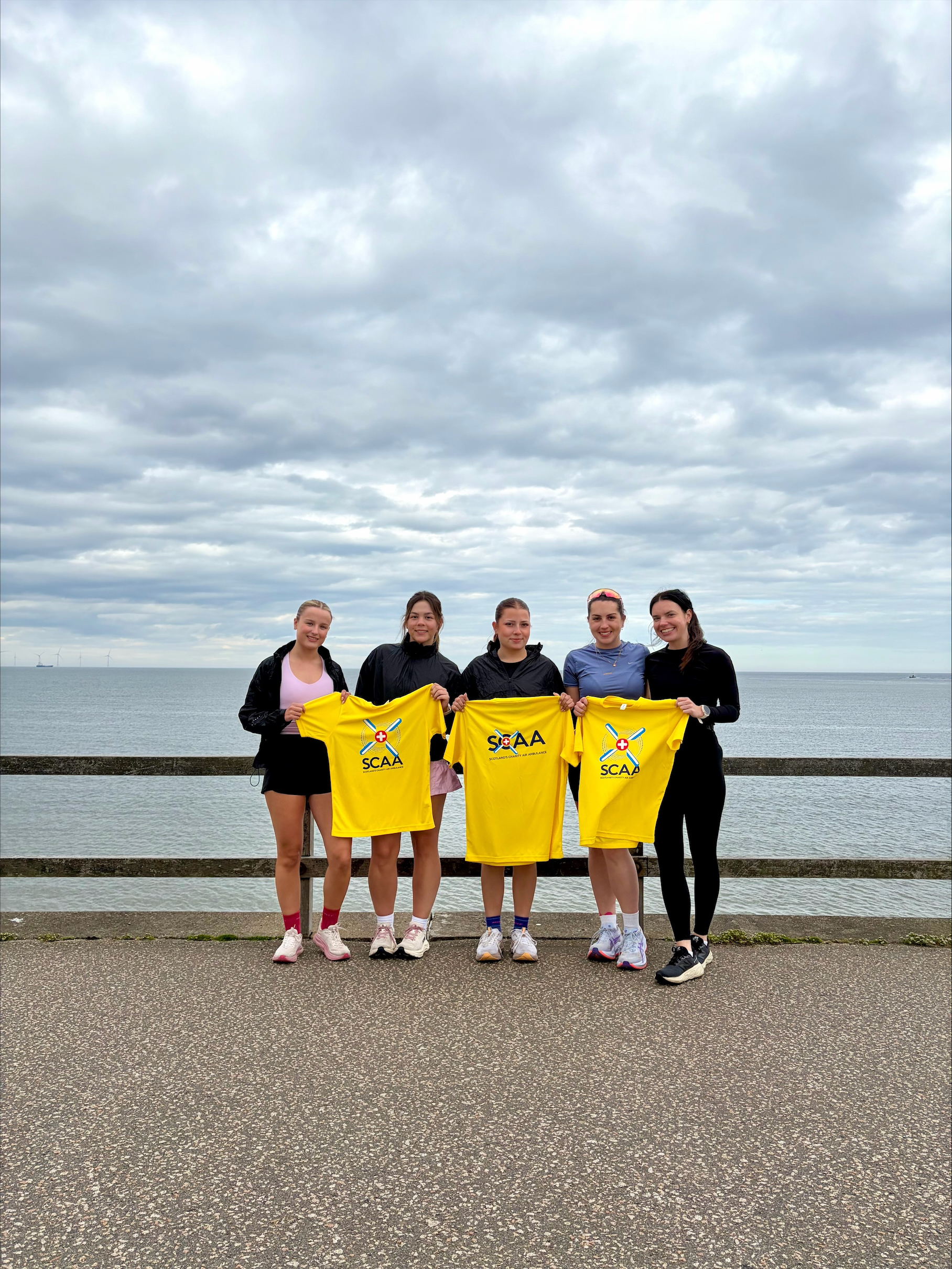 Five marathon runners each stand in front of a beach with grey clouds holding bright yellow shirts of the SCAA