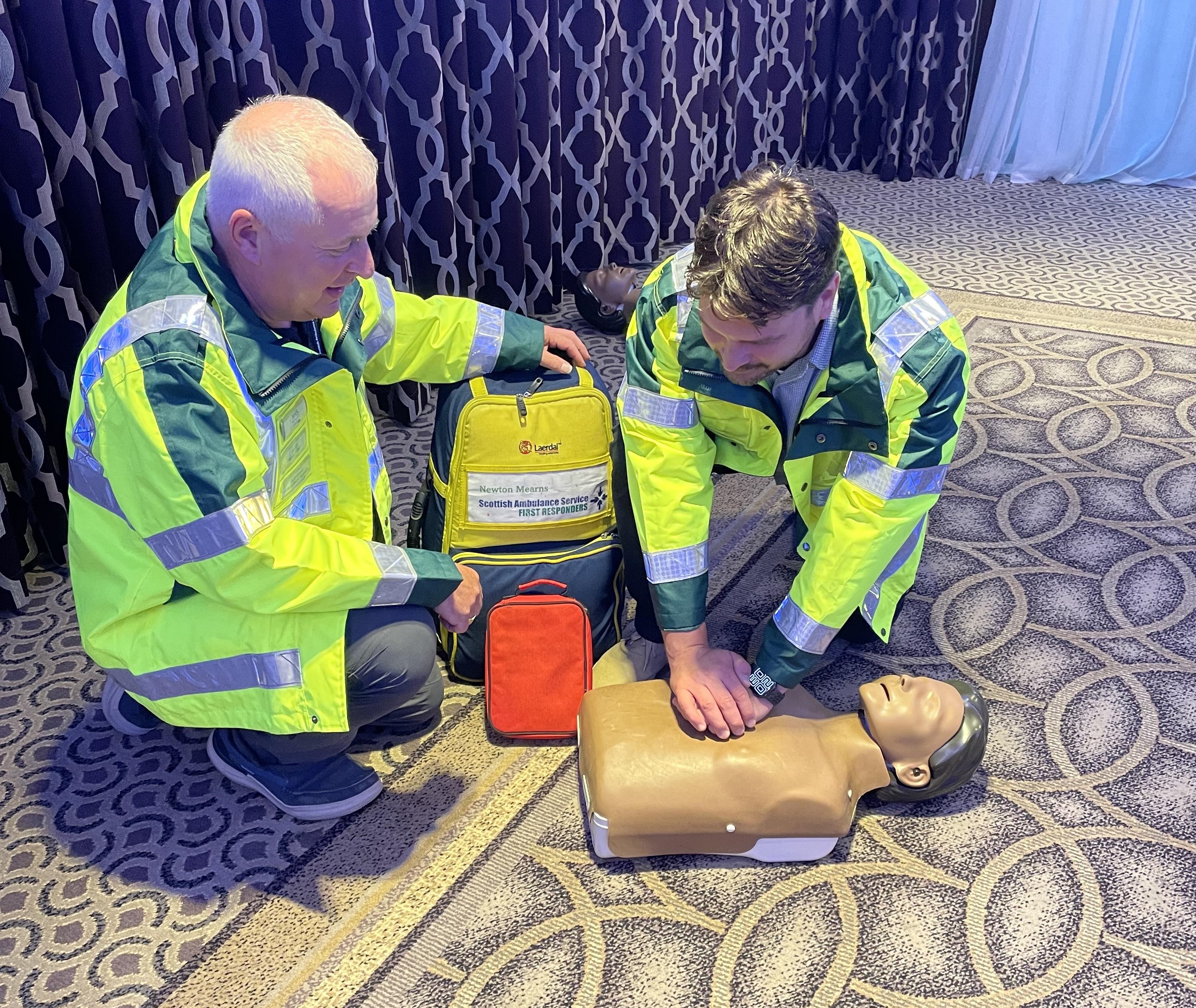 Two community first responders in high vis jackets demonstrate CPR on a CPR dummy,
