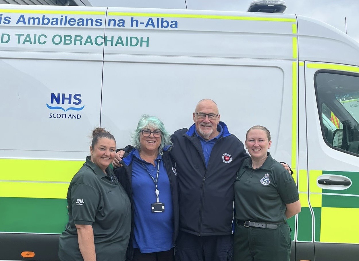 Sam Grieve, Mary and John Kinninmonth, and Laura Cameron standing together, smiling, in front of a parked ambulance vehicle