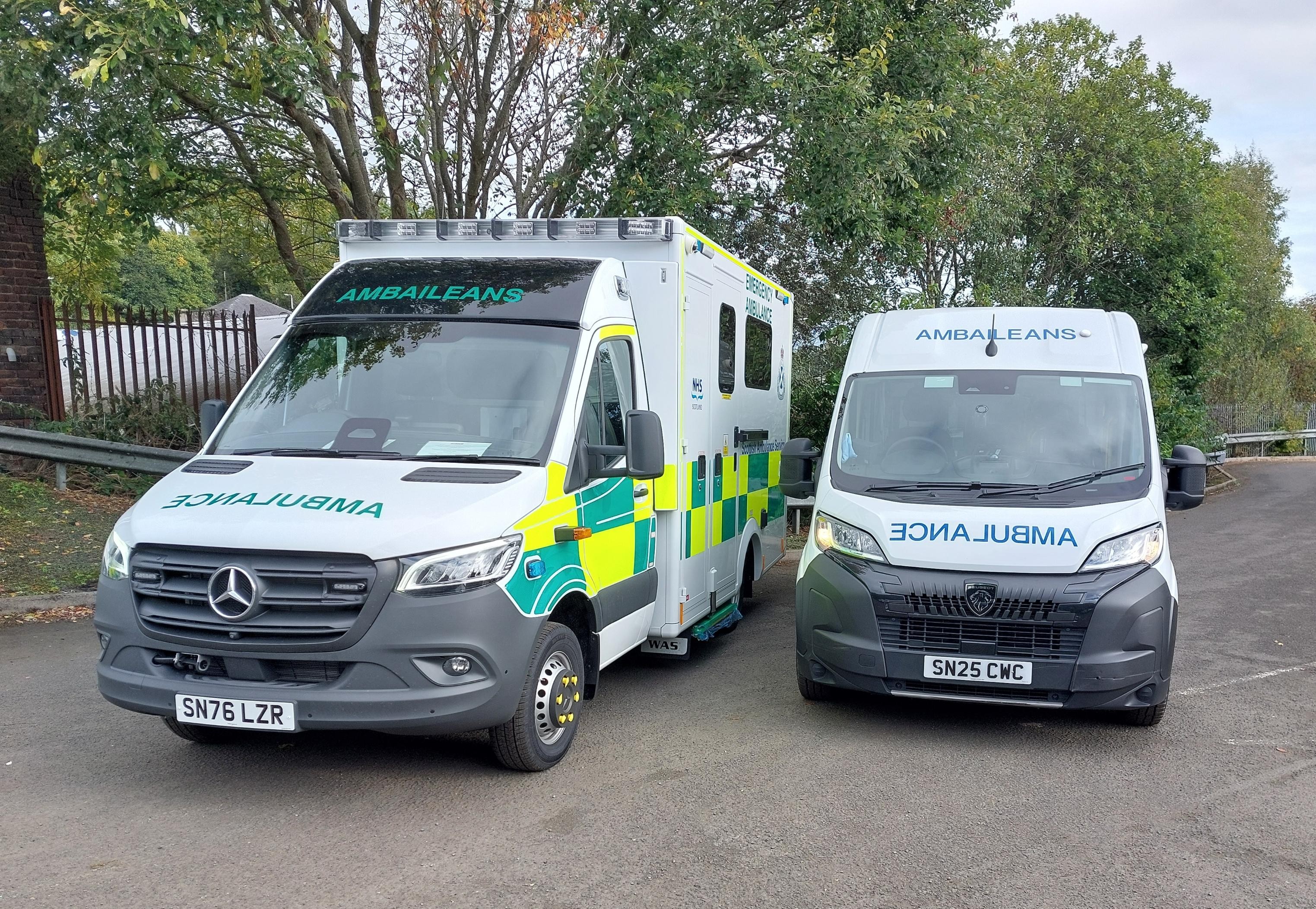 Two ambulance vehicles parked side by side on a paved area surrounded by trees and fencing.