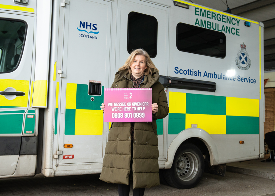 Lynsey Duncan holds a sign for CPR Bystander Support service whilst stood in front of an ambulance.