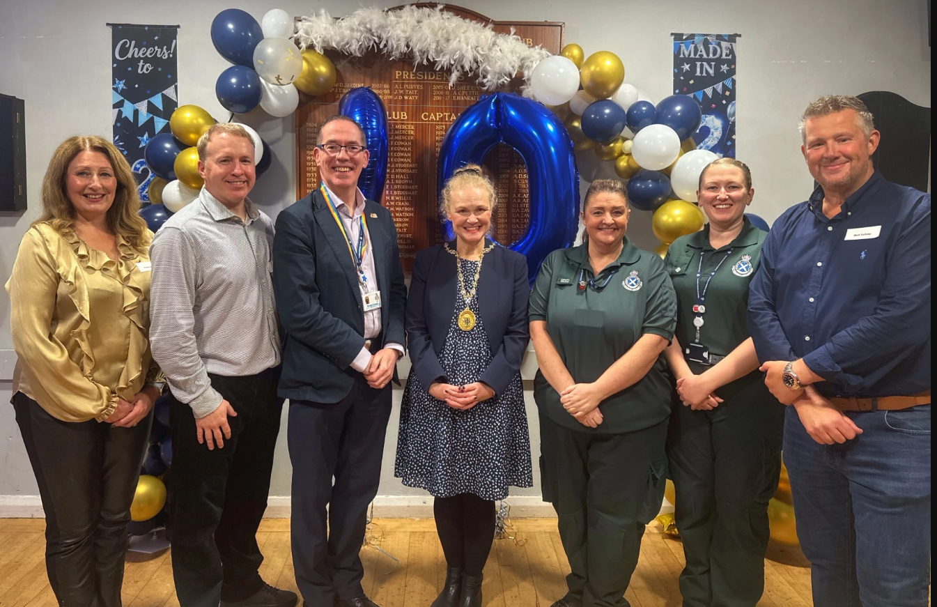 A group of seven people including Penicuik CFRs, SAS Chief Executive, stood together in front of balloons and celebration banners.