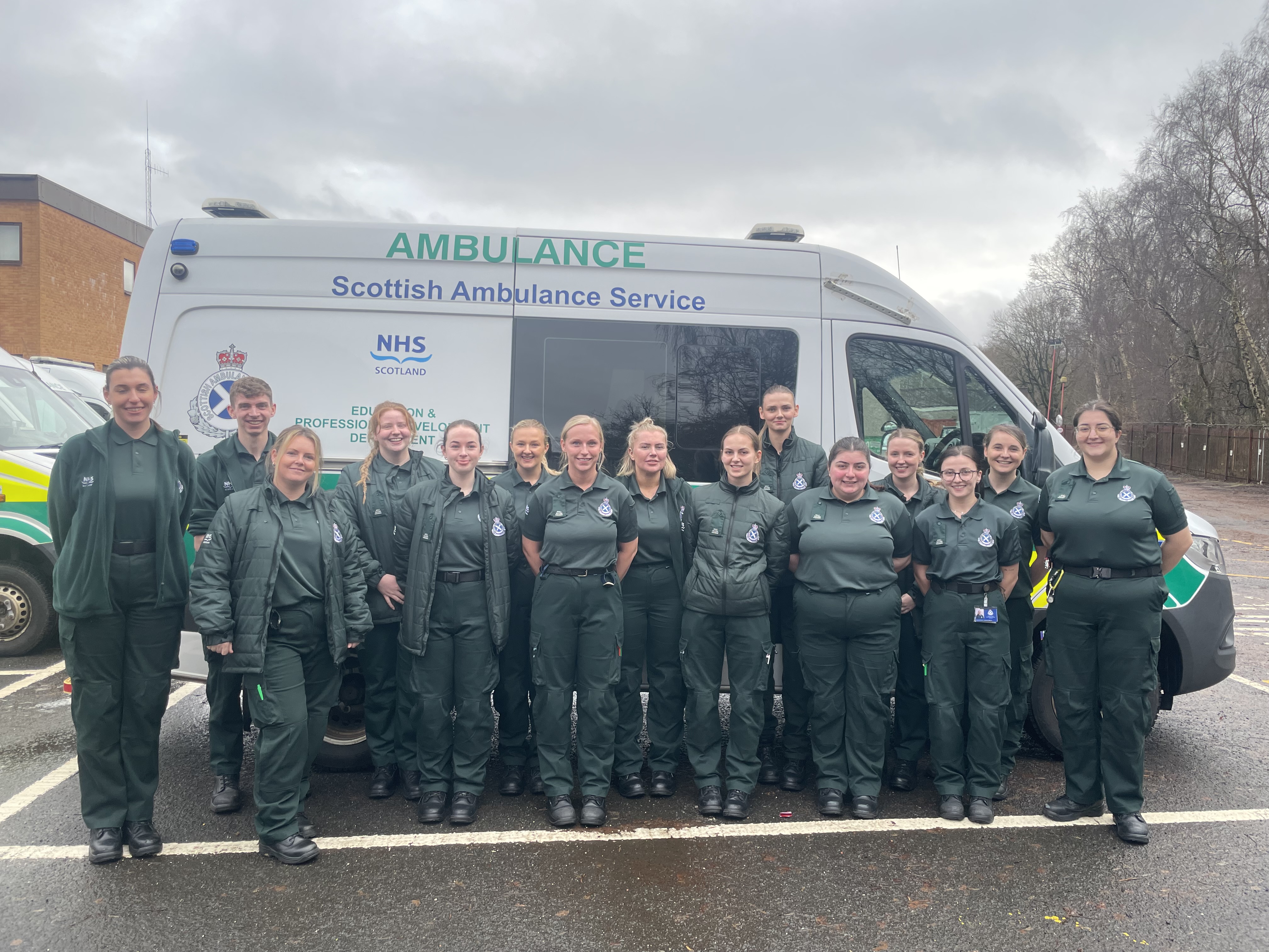A large group of newly qualified paramedics in dark green SAS uniform stood in front of a parked ambulance van.