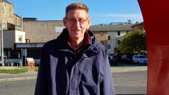 Man smiling at the camera on a sunny day behind him is a high street