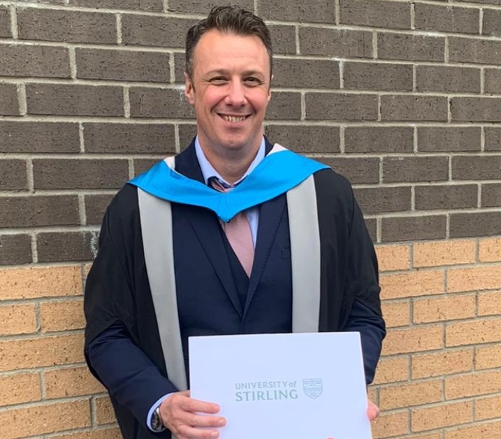 Dr Chris Aitchison wearing is graduation robes holds his certificate