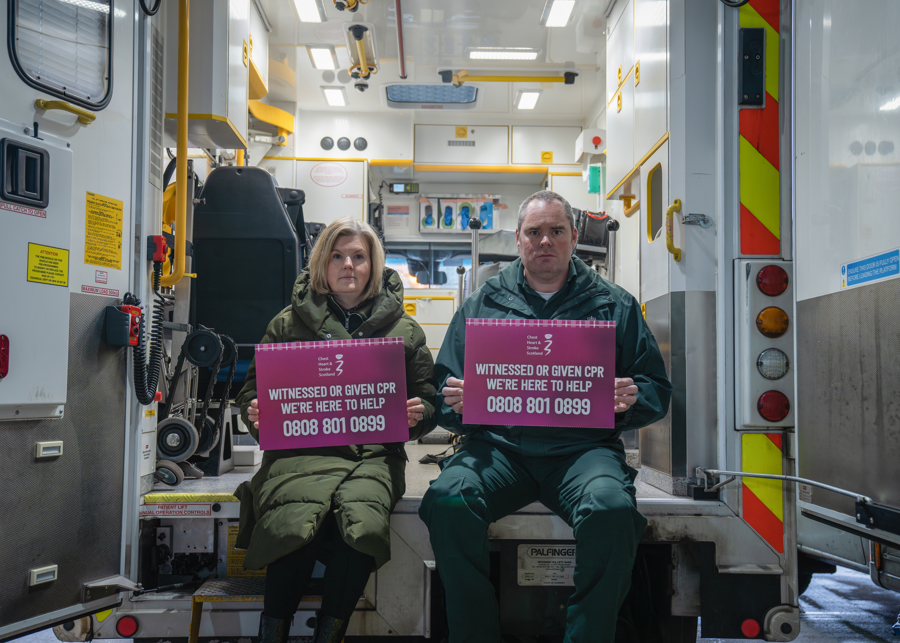 Lynsey Duncan and Steven Short hold signs for the CPR Bystander Support service whilst sat at the back of an ambulance with its doors open.