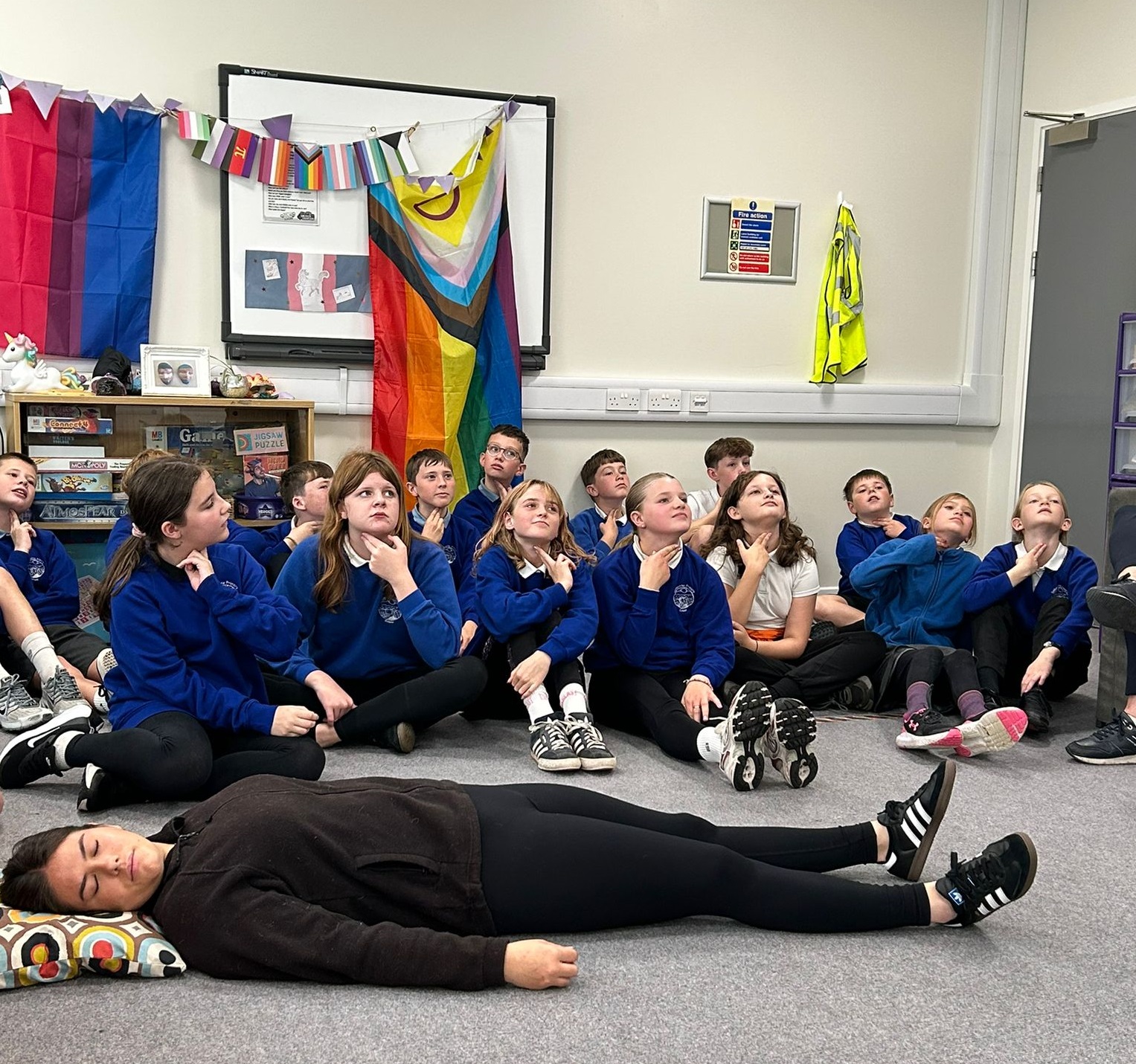 A woman laying on the floor pretending to be asleep in front of a full class of P7 pupils who are holding their hands to their throats to count their pulse.