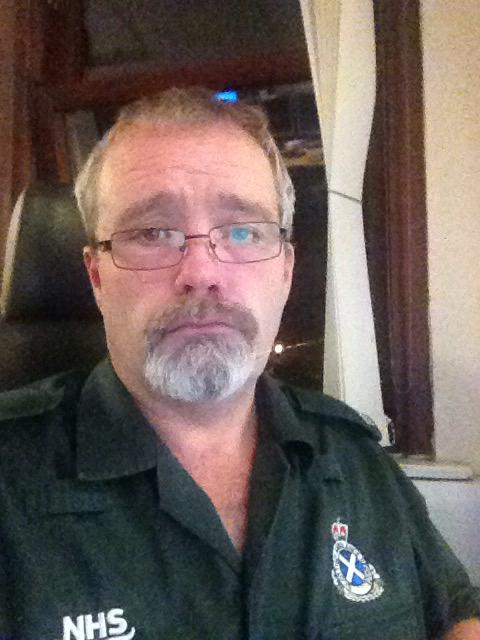 Calum sits at a desk expressionless in his green uniform 