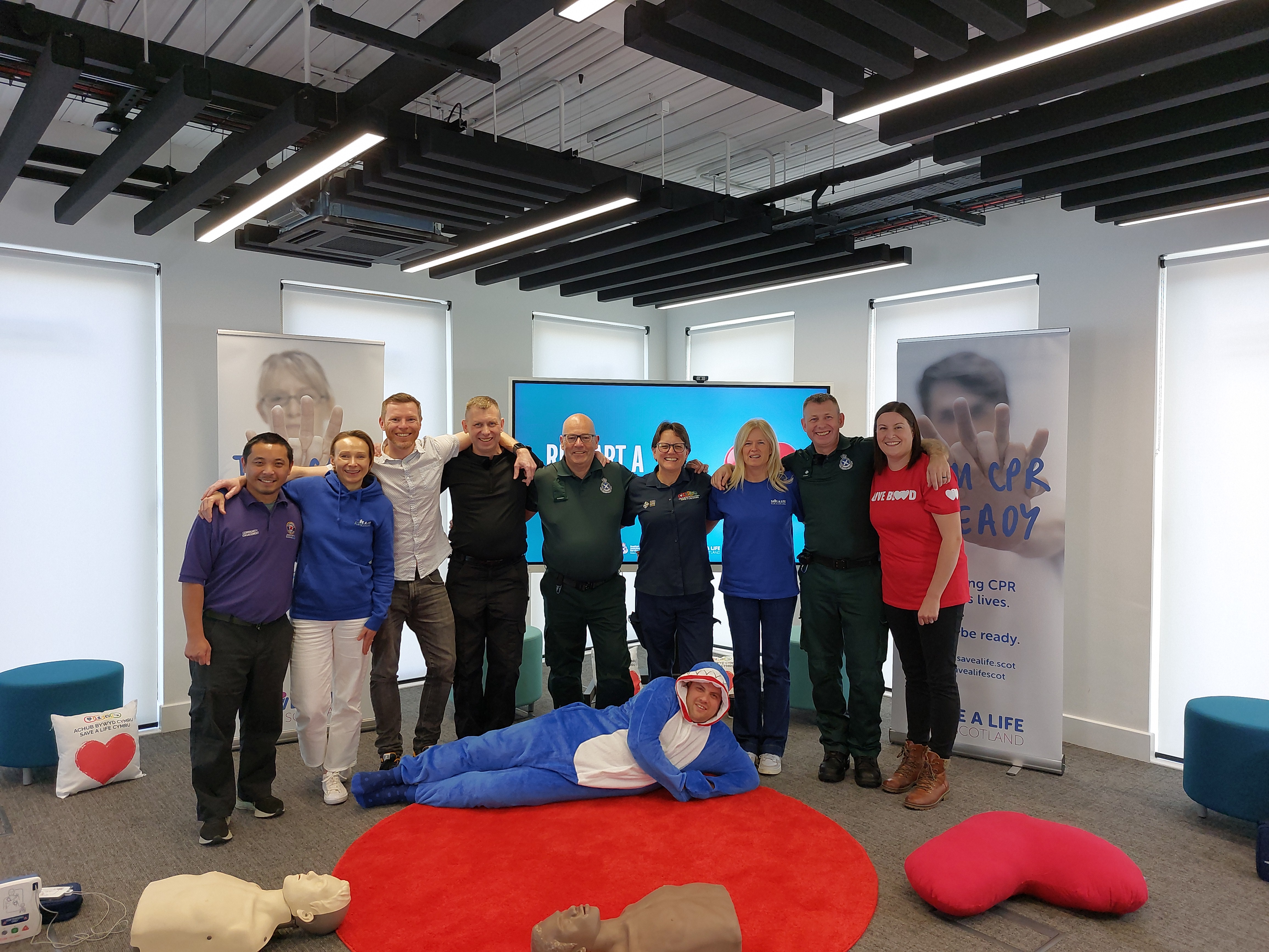 A group of 9 people standing together and smiling. Underneath them is a man laid on the floor in a shark costume.