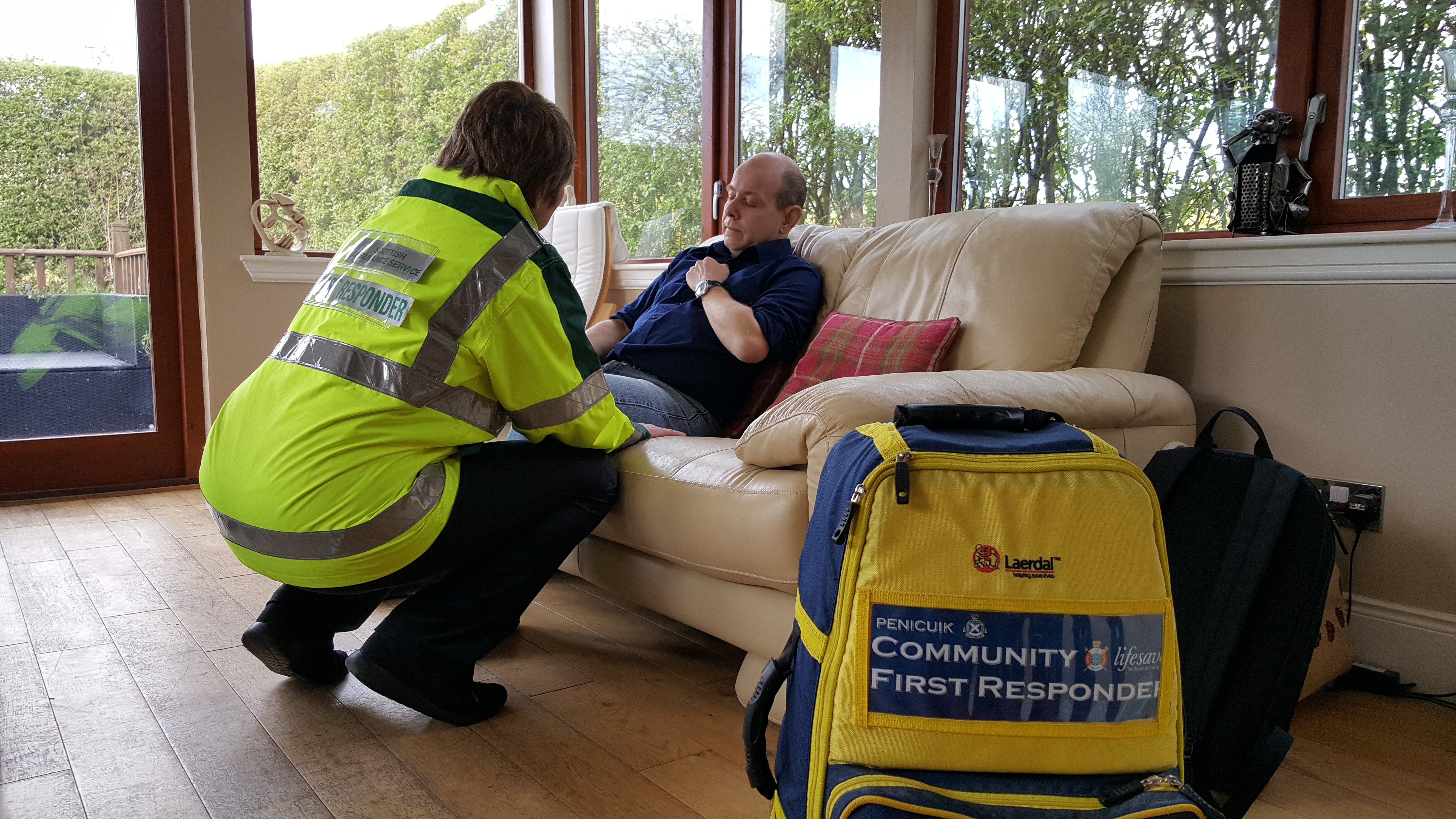 Female community first responder kneels while treating a male patient who is sat on a sofa.