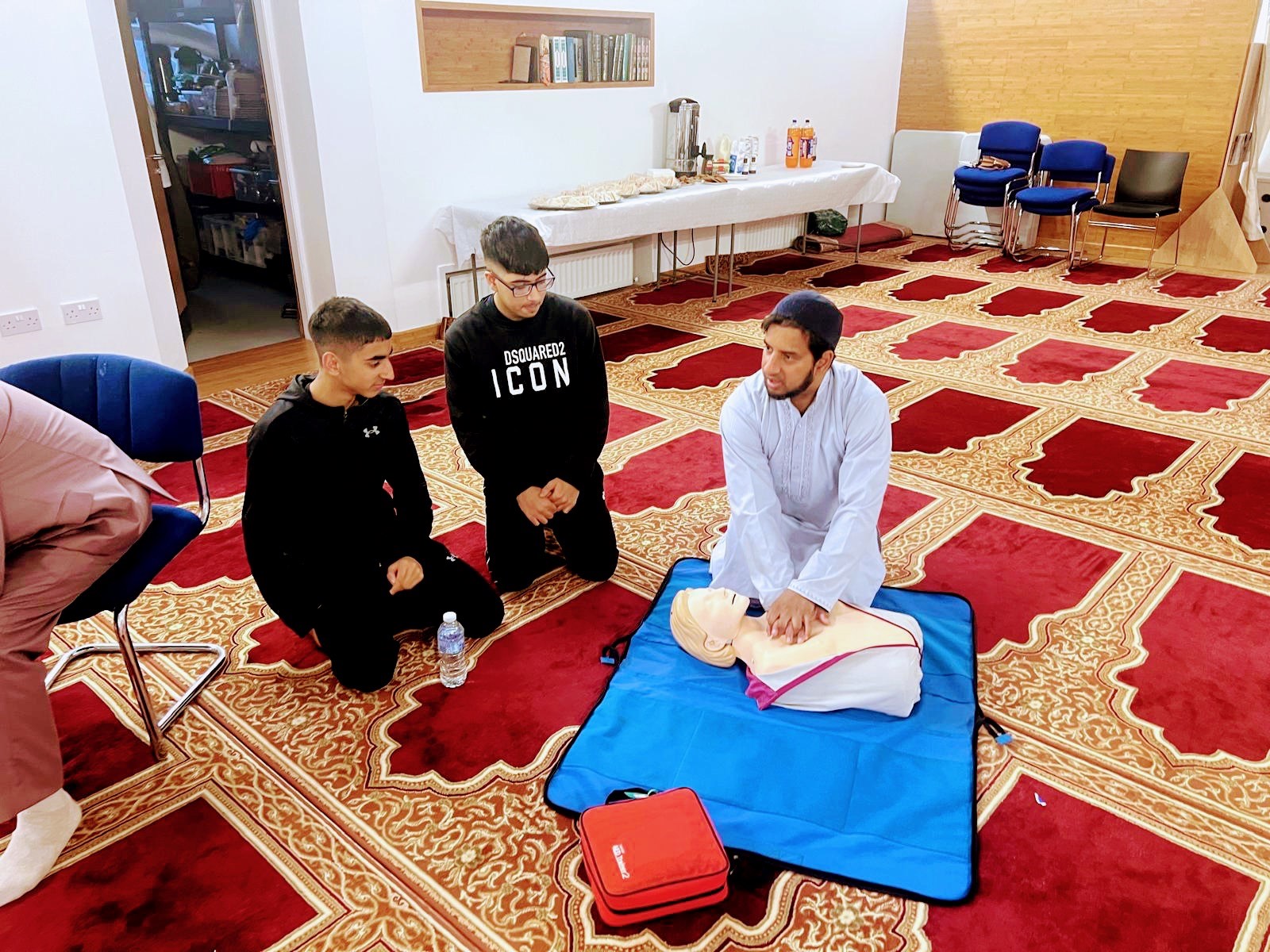 A man does CPR on a mannequin with two teenagers looking on 