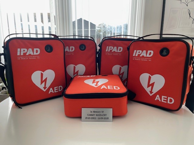 Five AEDs in red bags on a table. In front of them is a plaque that reads In memory of Cammy McGeachy, 19-10-2002 - 11-09-2025.