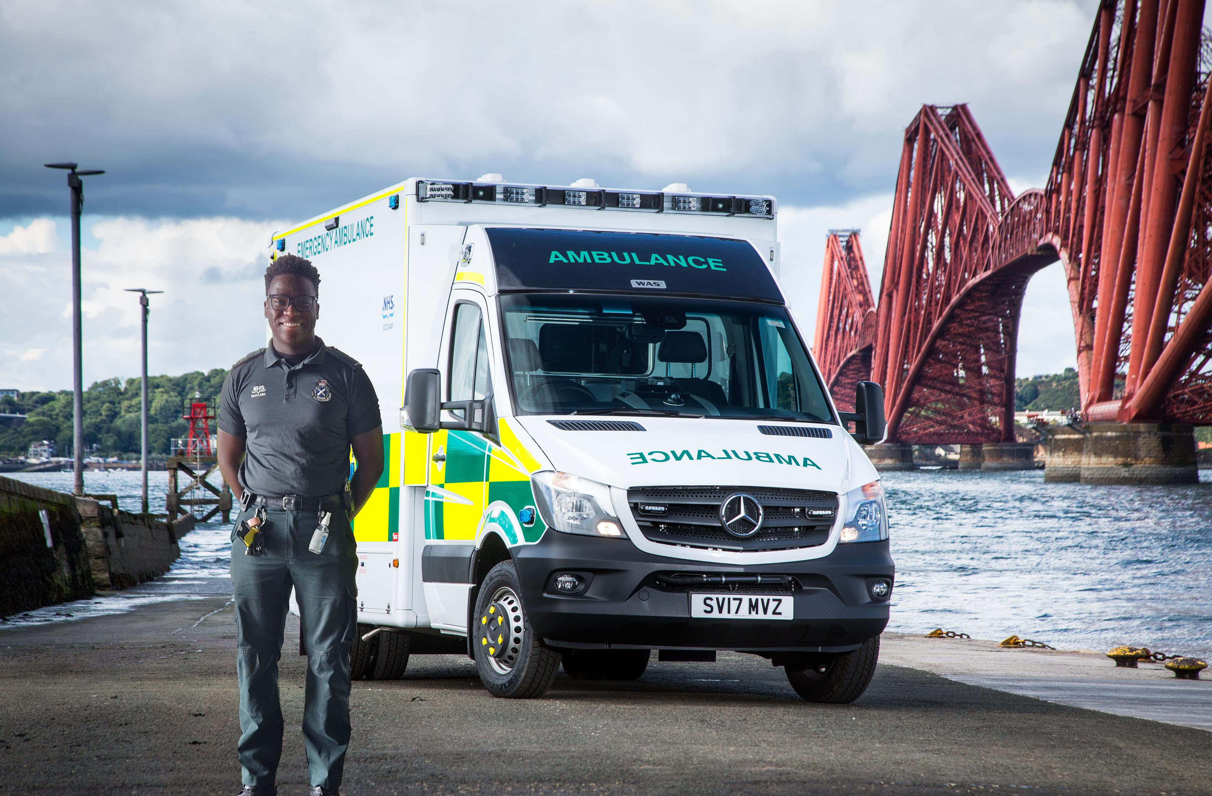An Ambulance Technician stands in front of an ambulance with the Forth Rail Bridge behind