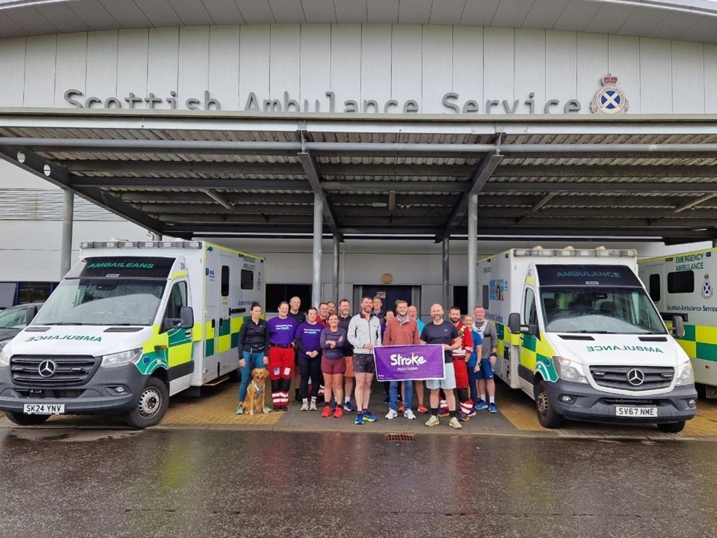 Scottish Ambulance Staff in front of an ambulance station between two ambulances holding the Stroke Association banner 