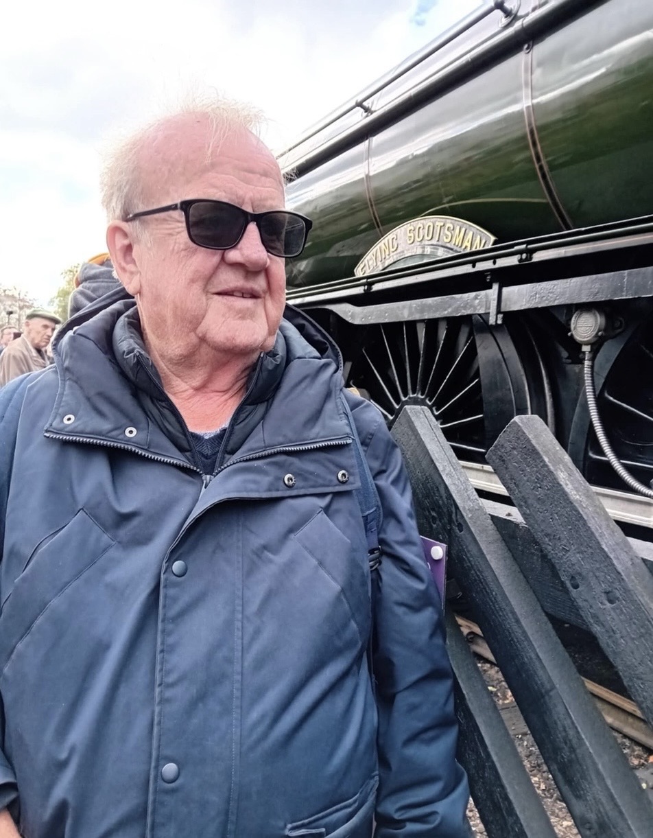 Jack stands in front of a train, which he loves, with glasses on