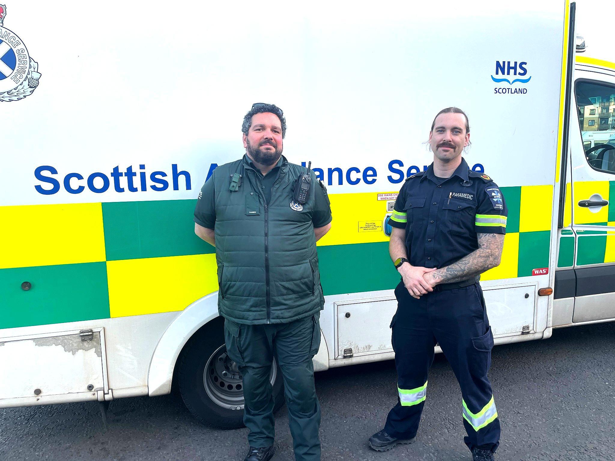 Paramedic Steve stands next to overseas staff member Luke in front of a Scottish Ambulance Service vehicle smiling