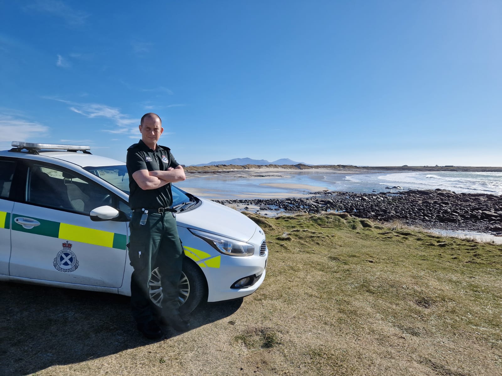 SAS Ambulance worker in uniform stood by a parked ambulance vehicle overlooking a beach