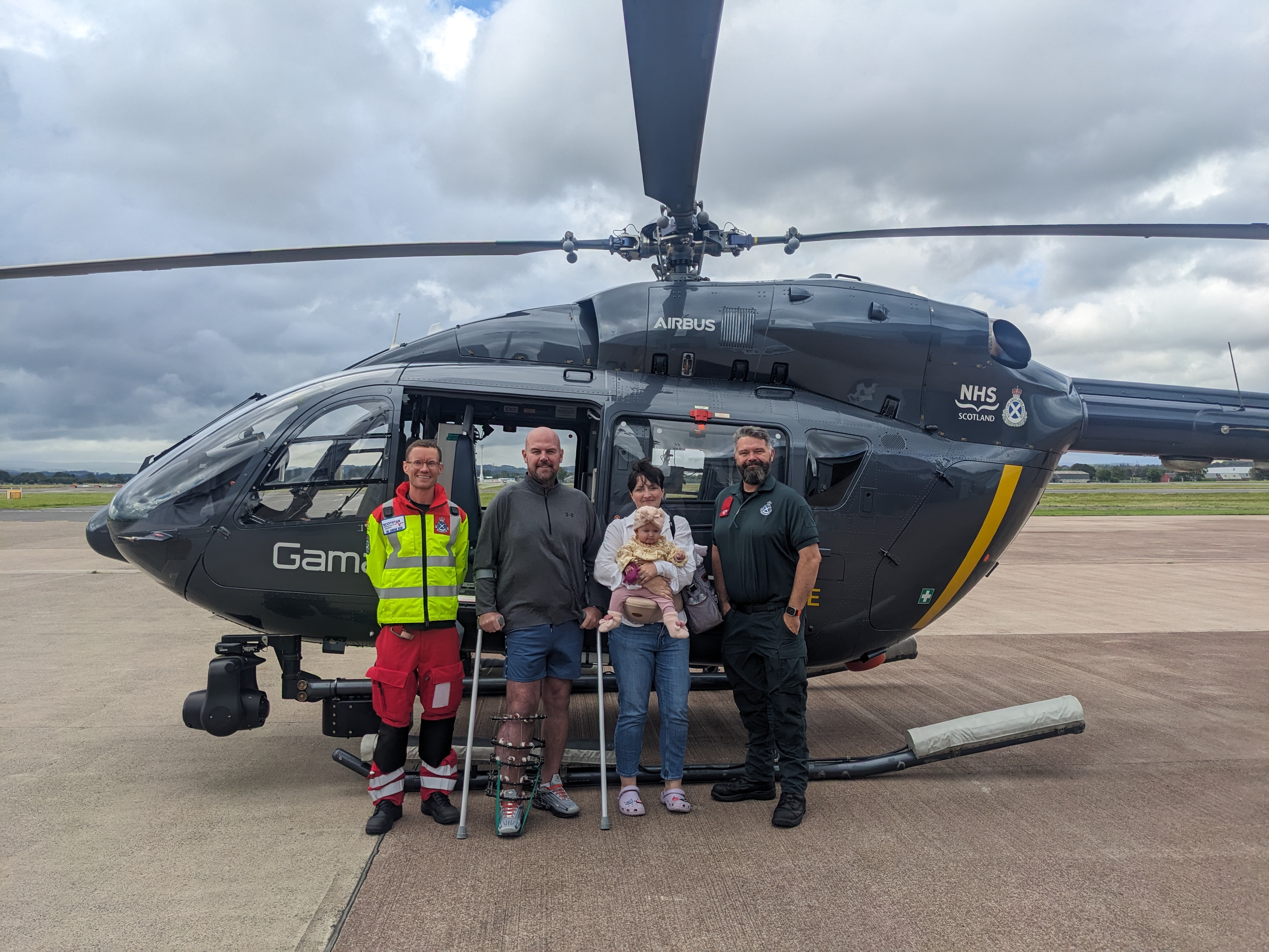 Four people and a baby stood together in front of a parked air ambulance helicopter on the tarmac at Glasgow Airport.
