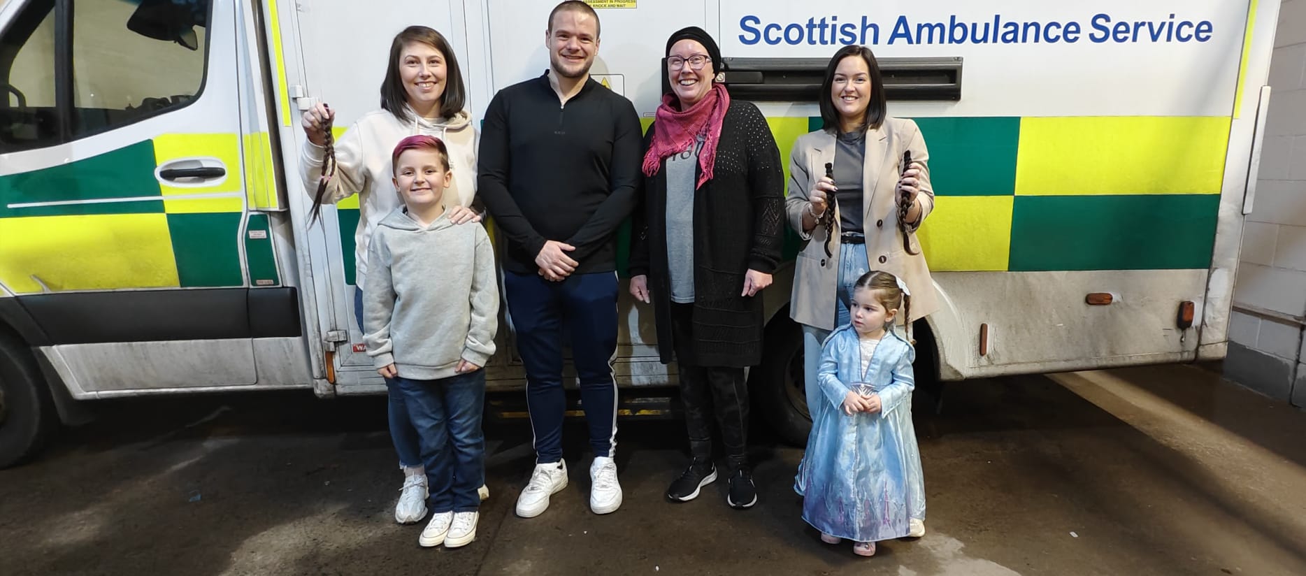 4 adults and two children standing together smiling in front of a parked ambulance.