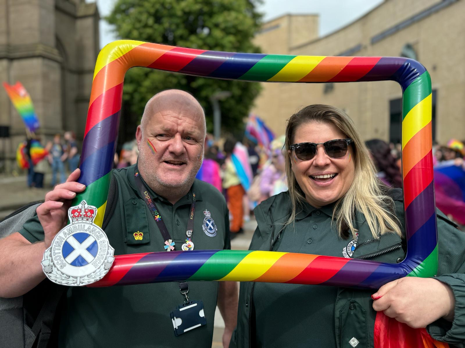 Two members of staff have their picture taken in a plastic rainbow frame