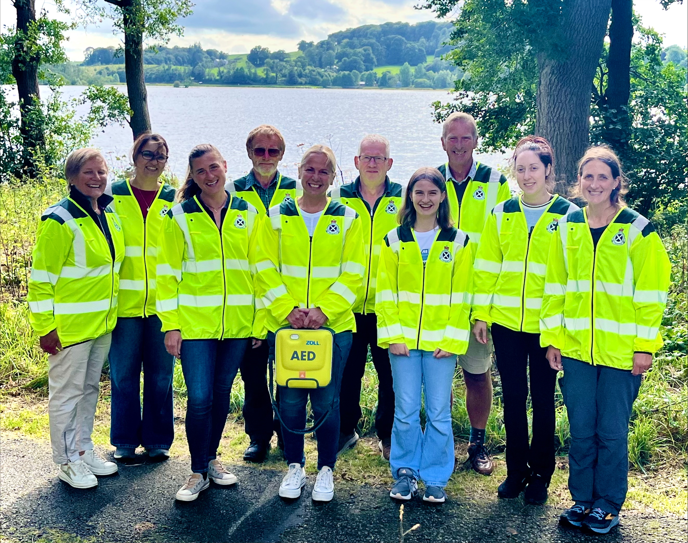 A group of 10 people in high vis jackets, one holding an AED, and smiling together